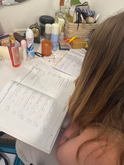An indoor scene depicts an individual seated at a cluttered white desk, with their back partially visible, facing away from the camera. The individual has long, light brown hair and a small, black maple leaf tattoo on their inner right arm. An open textbook, displaying Hebrew text and geometric problems (cubes, cones, cylinders) with multiple-choice options, lies in the foreground. Page 357 is visible. To the right of the textbook, an open spiral-bound notebook contains handwritten notes and diagrams in Hebrew.
The desk surface is crowded with various personal items. From left to right, these include a pink calculator, a clear bottle labeled "Aura," a small pink roll-on deodorant, a white and blue spray deodorant (Rexona brand), a brown object, an orange candle in a glass holder, a black elastic hair tie, a necklace, and a gold ring. Further back, an antique-style alarm clock, several tall cylindrical bottles (some clear glass containing liquid, others opaque white/light blue), and a dark container are arranged. A woven basket filled with assorted small items, including bags and containers, is also present. The background shows a light-colored wall, with a partial reflection from a mirror visible at the top edge of the image. The specific location within the Gilboa Regional Council, Israel, is not discernible from the image content.