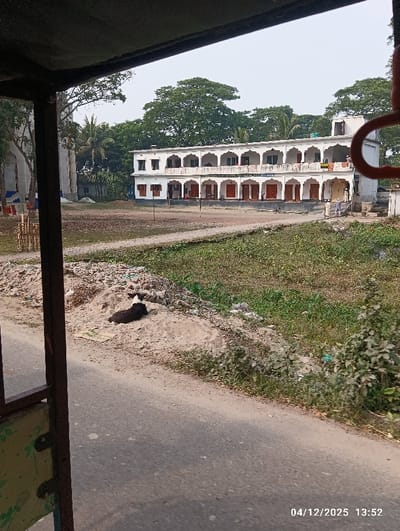 The image captures an outdoor scene in Kaliganj, Bangladesh, viewed from a vehicle frame, as indicated by the dark metal structure partially obscuring the top and left edges. The timestamp 04/12/2025 13:52 is visible in the lower right corner.
In the foreground, a paved road runs diagonally from the bottom left towards the center right. Adjacent to the road on the right, a black and white dog is resting on a dirt pile that contains scattered debris. Beyond the road and dirt pile, an unmaintained field of dirt and sparse grass extends towards the background; two vertical posts are visible in this field, suggesting a potential playing area.
The central background is dominated by a prominent two-story, white building featuring a facade with multiple arched verandahs on both floors. The ground floor displays several brown wooden doors, while the upper floor balconies are equipped with white railings. Several individuals are present near the building: two men in light-colored traditional clothing stand near the ground floor entrance on the right side of the building, and less distinct figures are visible on both the ground and upper levels. Laundry is hanging from a line on the building's right facade.
To the far left of the main building, a tall, light-colored structure with blue accents is partially visible, mostly obscured by trees. A dense canopy of large green trees forms the backdrop for the entire scene, under a clear, pale blue sky.