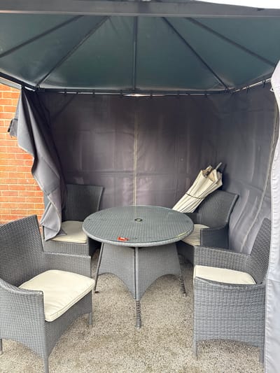 The scene depicts a garden gazebo containing outdoor furniture, located within the Metropolitan Borough of Solihull, United Kingdom. The gazebo features a dark grey fabric roof and side panels, supported by an internal black metal frame. One side panel on the left is partially drawn back, revealing a reddish-orange brick wall. Centrally positioned within the gazebo is a round, dark grey woven synthetic rattan table with a clear glass top and a central hole. Four matching dark grey woven synthetic rattan chairs, each fitted with a light cream-colored seat cushion, are arranged around the table. A folded, light beige patio umbrella is propped against the rear right fabric wall. A small, red and orange utility lighter or similar object rests on the glass tabletop. The ground surface within the gazebo consists of coarse grey gravel. No individuals are present in the scene.