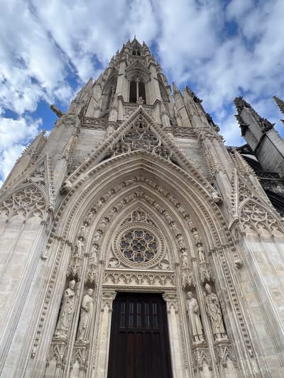 The image displays a detailed, upward-looking view of the Cathédrale Notre-Dame de Rouen (Rouen Cathedral) in Rouen, France. The central subject is the ornate Gothic stone architecture of the cathedral's facade, dominated by a tall, intricately carved central spire or pinnacle ascending towards the sky. Below the spire, a large, deeply recessed pointed arch portal forms the main entrance, featuring multiple layers of sculpted archivolts populated with numerous small figural statues. Above the dark, multi-paneled wooden doors at the base of the portal, a circular rose window with complex tracery is visible. Flanking the doors are several full-length stone statues in niches, including figures dressed in ecclesiastical robes, one holding a crozier and wearing a mitre. The stone work is light-colored, showing intricate details, pinnacles, and decorative tracery across the facade. At least two grotesque gargoyles project from the upper sections of the building. The background consists of a bright blue sky partially obscured by scattered white, fluffy clouds. Elements of a darker, perhaps older, section of the cathedral's structure are visible to the far right, contrasting slightly with the lighter main facade.