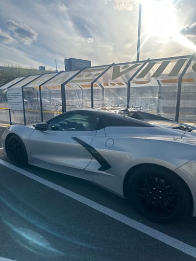 A light-colored (possibly silver or white) Chevrolet Corvette C8 sports car is parked in an outdoor parking area in Volketswil, Switzerland. The image is taken at a vertical orientation, showing the bright, sun-drenched sky and clouds on the left, and the asphalt parking lot on the right. The sun is high in the sky, indicating bright daylight, likely late morning or afternoon, and casts strong glare and reflections.
The car is positioned next to a translucent shelter-like structure with yellow-patterned panels. Parking space lines are visible on the asphalt. To the left of the car, beyond the shelter, are what appear to be shopping carts. On a sign attached to the shelter, German text is visible, stating "VOLKILAND" at the top. The sign also reads: "Die Nutzung des Parkareals ist nur während der Öffnungszeiten von 8 bis 20 Uhr gestattet. Bitte nehmen Sie Rücksicht auf die Anwohner. Vielen Dank für Ihre Unterstützung." (The use of the parking area is only permitted during opening hours from 8 AM to 8 PM. Please show consideration for the residents. Thank you for your support.)
In the background, a distant building with the "weber" logo can be seen, along with other industrial or commercial structures and a tall smokestack or chimney under the partly cloudy sky. The car features black multi-spoke wheels, and the sleek lines of its body are highlighted by the bright sunlight. No people are visible in the scene.
