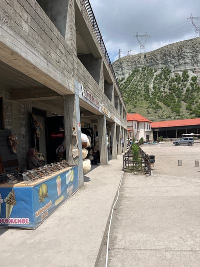 The image depicts a street scene in Chirkey, Russia. A multi-story, unfinished concrete building dominates the left side of the frame, its raw construction visible with open window spaces. At ground level, a shop is open, displaying various items including stuffed animals and souvenirs. An ice cream advertisement with Russian text is positioned at the edge of the shop's storefront. Higher up on the building's floors, there are displays of sheepskin hats or similar items.
In the background, there is a mountain covered with vegetation and power lines crossing the sky. Further back, there is a paved area with a building featuring a red roof and at least one vehicle.
The ground is concrete. A white cable runs along the ground.