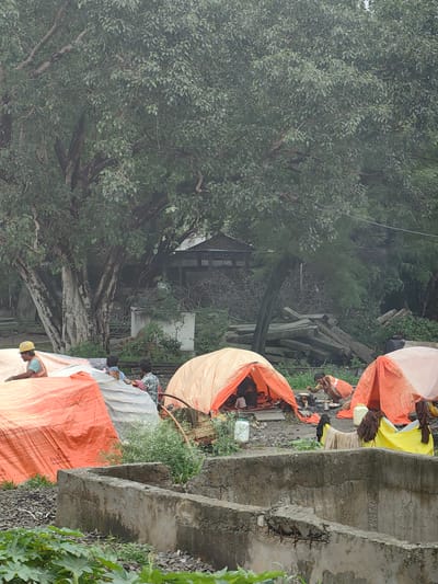 The image depicts an outdoor scene likely in Bhopal, India, characterized by the presence of temporary shelters and debris. In the foreground, a partially constructed concrete structure is visible, with exposed rebar suggesting ongoing or abandoned construction. Beyond this, several tents, primarily orange and white, are erected, serving as makeshift shelters.

Several individuals are present. On the left, a person wearing a yellow hard hat and an orange vest is partially visible, interacting with materials covered by orange tarpaulin. In the center, within an orange tent, two individuals are seated. To the right of this tent, a woman is crouched down, appearing to be cooking or preparing food. Further to the right, another orange tent is set up, with clothes hanging to dry on a yellow tarp nearby. Scattered around the tents are various objects, including what appear to be water containers and a pile of concrete beams in the background.

The environment is dominated by a large, leafy tree in the middle ground, with dense foliage suggesting a humid climate. Behind the tree, structures with corrugated metal roofs are visible, indicating a more permanent settlement or industrial area. The ground is covered with gravel and dirt, and there is vegetation growing around the concrete structure and between the tents. The overall impression is one of temporary living arrangements amidst a backdrop of construction and natural elements.