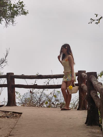 A woman stands on a lookout point in Hải Phòng, Vietnam. The sky is overcast. She is fair-skinned, with long brown hair adorned with a starfish clip. The woman wears a light yellow tank top, a floral skirt, and flip-flops, and carries a small straw bag decorated with a yellow flower. She is touching her hair with her left hand. The lookout has a low, wooden railing with shrubs and trees growing beyond. A concrete path leads to the lookout point.