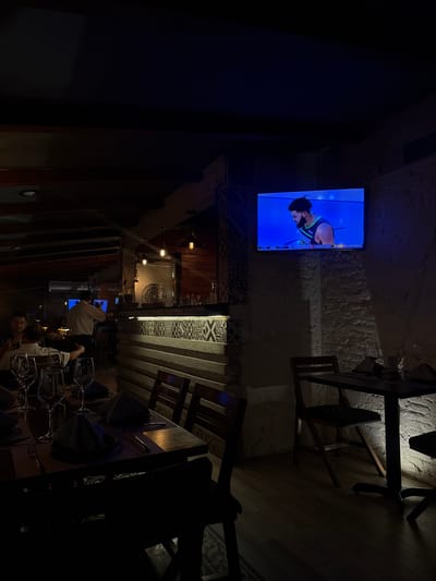 Here is a detailed description of the image:
**Scene Overview:**
The image appears to be taken inside a dimly lit restaurant or bar in Tuxtla Gutiérrez, Mexico. The view is from a high angle, possibly from a mezzanine or elevated seating area. The lighting is focused on specific areas, creating a contrast between light and shadow.
**Subjects & People:**
* There are a few patrons and staff visible in the lower part of the image, near tables set for dining. They are wearing white and dark shirts, and appear to be interacting with each other.
**Objects:**
* **Tables:** Several dining tables with silverware and glasses are arranged in the lower part of the image.
* **Chairs:** Chairs are positioned around the tables.
* **Television:** A television is mounted on the wall, displaying a sports game or program with a basketball player.
* **Lighting Fixtures:** Wall-mounted lighting fixtures provide some illumination.
* **Bar Area:** There's a bar area in the background with bottles and bar equipment.
* **Architectural elements:** Various design elements can be seen, including decorative wood panels and textured walls.
**Actions & Interactions:**
* Patrons and staff seem to be engaged in conversation and dining.
* People are watching the television.
**Environment:**
* The restaurant has a mix of modern and rustic design elements.
* The ambiance seems relaxed and casual.
* There is a long staircase on the left side of the frame.
**Location:**
* Tuxtla Gutiérrez, Mexico is the city, according to the prompt. It seems to be a local restaurant or bar.