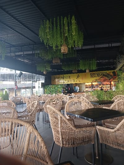 The image shows a restaurant patio with wicker chairs and tables, located at the coordinates -1.109 latitude and 36.71 longitude in Riara Ridge, Kenya. The patio is covered by a black metal awning with hanging green plants. The restaurant is adorned with a sign reading "JOHNNIE WALKER" and "KEEP WALKING". The chairs and tables are arranged in rows, with the chairs facing the tables. There are a few people in the background, sitting at the tables. The image is taken from a low angle, looking up at the patio.