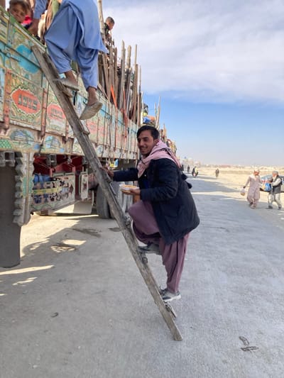 An outdoor scene in Chaman City Tehsil, Pakistan, depicts a large, ornately decorated truck and several individuals on a paved surface adjacent to a dusty, open landscape under a partly cloudy sky.
The truck, brightly painted in blues, greens, and reds with geometric and floral patterns, and the word "ANYS" visible on its side, is stationary on the left. A rustic wooden ladder leans against the truck's side. An individual, wearing a light blue shalwar kameez and light-colored slip-on shoes, is seen ascending the upper portion of the ladder, with their back partially to the camera.
Below this individual, a young adult male stands on the lower portion of the ladder, facing the camera and smiling. He wears a dark blue jacket over a dark undershirt, maroon shalwar kameez, and a light pink patterned scarf wrapped around his neck. His feet are clad in grey and white athletic shoes. He holds a small, light-colored bowl containing yellowish food in his left hand, and his right hand rests on the ladder. Within the truck bed, several individuals, including children, are partially visible among various wooden poles and cargo. One child in the upper left corner looks towards the camera.
The foreground consists of a grey paved surface, exhibiting shadows from the truck and individuals. The background to the right reveals a wide, dusty, unpaved area extending to a hazy horizon. Several figures are visible in the mid-ground: two individuals in light-colored traditional attire, one carrying a white bag, and a light-colored vehicle partially visible behind them.