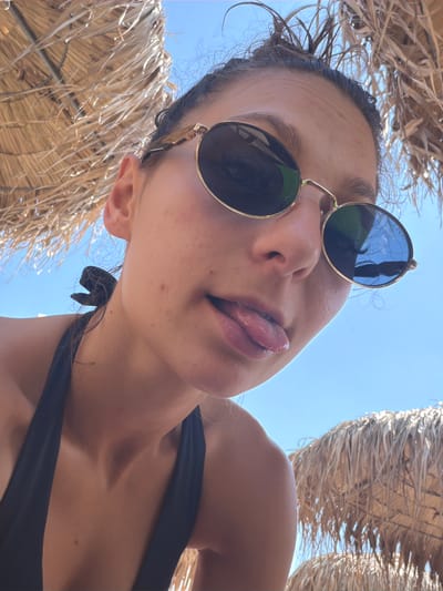 The image is a low-angle, close-up shot of a young woman from the chest up, outdoors under a thatched umbrella against a bright blue sky. She is wearing a black bikini top and gold-rimmed sunglasses. Her hair is pulled up, with some strands falling loose. She is sticking her tongue out slightly. The background consists of the straw of the umbrella and the clear sky, with another thatched umbrella visible at the bottom right. There are no discernable features indicating the location is the city of Plovdiv, Bulgaria.