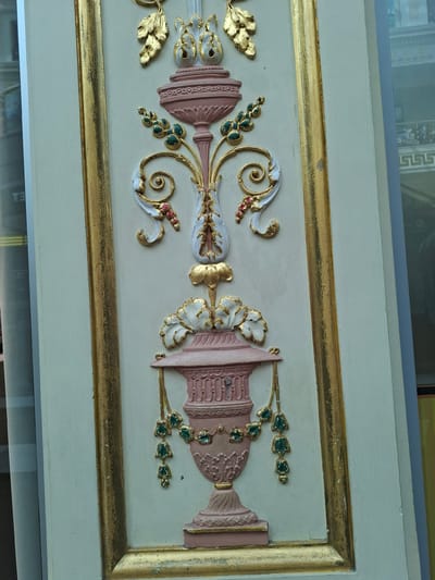 The image shows a close-up of a decorative relief panel, likely part of an architectural element or furniture. The panel features an ornate design with a central vertical motif resembling a stylized vase or urn. This motif is rendered in shades of pink, cream, and gold, with accents of green and red. There are floral and foliate elements, scrolls, and what appear to be beads or berries. The panel is framed by a wide, textured gold border. The background of the panel is a pale cream color. Reflected in the surface to the left of the panel, and in the background to the right, are elements of an urban environment, including buildings and what appears to be a shop or interior space. There are no people visible in the immediate foreground of the image. The image is taken at an angle, and the lighting suggests it is daytime. There is no specific indication of its location within the city of Trafford, United Kingdom, but the architectural style and decorative elements might suggest a public building, a historic property, or a shop interior.
