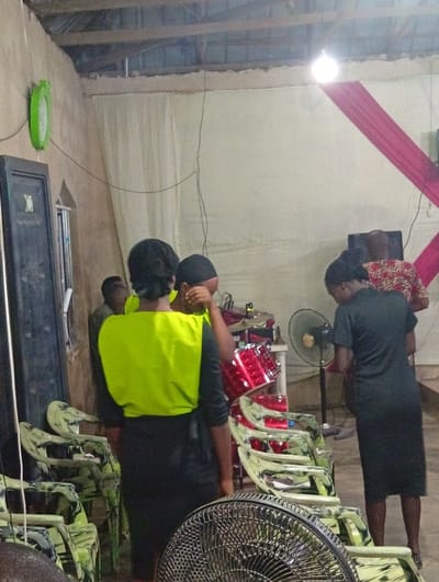 This indoor photograph depicts a scene within what appears to be a community hall or a simple church building in Nando-Ikem, Nigeria. Several individuals are present, engaged in what seems to be preparation or informal gathering.
In the foreground center, a person is seen from the back, wearing a black dress beneath a bright neon green sleeveless top or vest. To their right, another person in a dark, short-sleeved dress is also facing away. Further right, a person in a patterned shirt is partially visible. On the left, a man in a green shirt is partly obscured.
The room is furnished with numerous plastic chairs featuring a distinctive green and black camouflage-like pattern, some arranged and others stacked. A prominent red drum kit stands in the mid-ground on the right, suggesting a musical event or practice. Two electric fans are visible: one floor-standing fan near the drums and the top of another fan in the immediate foreground, bottom right.
The interior features exposed wooden roof trusses and ceiling material, indicating a basic construction. The walls are covered with light-colored fabric drapes, with a diagonal section of pink fabric on the right wall. A bright, bare light bulb hangs from the ceiling, providing primary illumination. A green oval-shaped wall clock is mounted on the left wall. Various electrical wires run along the walls and ceiling.
The overall impression is that of a local community or religious gathering space, possibly preparing for or winding down from an event involving music. The lighting suggests it might be evening or an enclosed space. No visible text is discernible in the image.