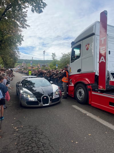 An outdoor scene in Molsheim, France, depicts a black and chrome two-tone Bugatti Veyron 16.4 supercar on an asphalt road, facing slightly left from the viewer's perspective. The vehicle bears a front license plate reading "BUG III". To its right, a white and red DAF 1045 truck is partially visible, featuring a red fairing with "TEAM" written vertically and an "ALSACE" shield emblem on its cab side. A male event staff member, wearing a high-visibility orange vest with "SCA" visible and a dark cap, stands between the Bugatti and the truck, facing the Bugatti. A dense crowd of spectators, many holding mobile phones, lines the left side of the road behind metal barriers, observing the vehicles. In the background, behind the crowd, a historic stone building, possibly a fortified gate, is visible, leading to a landscape of buildings, vineyards, and green hills under a partly cloudy sky. Mature trees line the left side of the road, and a vintage-style street lamp is present in the mid-background. The Bugatti appears to be stationary or moving slowly, with the staff member positioned as if managing traffic or observation.