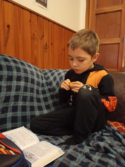 In this image, a young boy is sitting on a couch covered with a blue and gray plaid blanket. He is wearing a black hoodie with orange accents and black pants. The boy is looking down at something in his hands, which appear to be a piece of food, possibly a tangerine segment. He is intently focused on this activity.
To his left, an open book lies on the blanket. The book has text and illustrations, suggesting the boy might be reading or taking a break from reading. The background features a wooden paneled wall, indicating an indoor setting. Part of a wooden door is also visible on the right side of the frame.
The lighting suggests it is daytime, possibly late afternoon or evening, given the indoor setting and the soft lighting. The overall mood appears calm and domestic, with the boy engaged in a quiet, solitary activity.
Visible text on the book includes Cyrillic characters, such as "Снеговики", "Морковкин", "Варежкин", and what seems to be a title "KRATУНКОВ И КастрюлARRNOR". The text appears to be from a children's book.