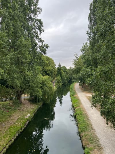 The image depicts a view from an elevated position looking down at a canal in Villeparisis, France. Lush green trees line both sides of the canal, with the left bank featuring a grassy area and a path, while the right bank has a wider, gravel path. The canal water is dark and reflects the overcast sky and the surrounding foliage. In the distance, the canal curves to the left, bordered by more trees. The scene suggests a serene, natural setting within or bordering the city. There are no visible people in the frame.