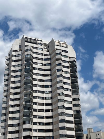A tall, modern apartment building is captured from a low angle against a bright blue sky filled with white, puffy clouds. The building, located in Créteil, France, features a distinctive facade with alternating bands of light beige and white, creating a zigzag pattern up its many stories. Each floor is lined with numerous windows and balconies, many of which have dark tinted glass or visible curtains. The building's architecture is angular and geometric, with pointed elements at the roofline. The image appears to be a photograph taken on a clear day, emphasizing the verticality of the structure against the expansive sky. No people are visible in the frame.