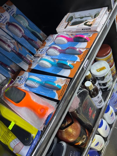 This image depicts a retail shelving unit inside a commercial establishment in Alasia, Nigeria, displaying various beauty and personal care items. The shelves are constructed of metal, possibly chrome. The primary focus is on multiple "4 in 1 Pedicure Paddle" sets, each packaged in clear blister packs on cardboard backings. These paddles feature colored handles in pink, purple, teal, orange, and black, incorporating a foot brush, pumice stone, emery file, and heel rasp.
On an upper shelf, a white rectangular box containing a "FLFWLASS FACIAL ROLLER & MASSAGER" with a jade roller is visible. Below and to the right, a collection of bottled and jarred products is arranged. This includes several dark brown bottles with gold caps labeled "Binuru Buba Rose Water," stacked white jars with green/yellow labels, and a row of white and blue Nivea brand lotions or body washes. A dark-colored bottle featuring a pink rose graphic and the brand name "IMAN" is also present.
A white price tag displaying "4,800" is affixed to a vertical shelf support, and a yellow sticker with "780" is visible on the packaging of a black-handled foot care product. No individuals are present in the scene, and no actions or interactions are depicted. The lighting suggests an indoor retail environment.
