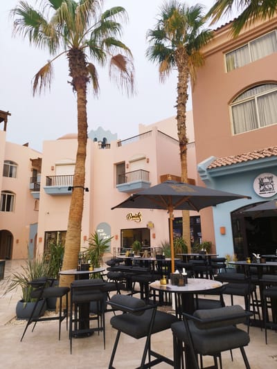 This image shows an outdoor café or restaurant in Ras Abu Soma, Egypt, under an overcast sky. The space is furnished with numerous small, round tables and matching dark-colored high chairs. Several tables are adorned with condiments. Umbrellas provide shade to the tables. The surrounding architecture consists of low-rise buildings, primarily in shades of peach and light blue, with some balconies and arched windows. Tall palm trees stand between the seating area and the buildings. Signs for “Breads” and “House of Seven” are visible above the entrances to businesses. Plants in pots add greenery to the scene. The ground is paved with light-colored stones. No people are visible.