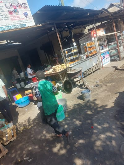 The image depicts a street scene in Picha Ya Ndege, Tanzania. In the foreground, a young boy wearing a green shirt with a black and white pattern is walking away from the camera. To his left, a light blue plastic basin and a green plastic container are visible on the ground. Behind the boy, a man in a white shirt is seen interacting with a woman in a black and white striped apron, who is standing near several white buckets. Further back, a market stall with a corrugated metal roof is set up, displaying food items in glass cases. A sign with text in Swahili is visible on the left side of the image. The ground is dirt and appears to be a public space, possibly a marketplace. The overall impression is that of a busy, everyday scene in a Tanzanian town.