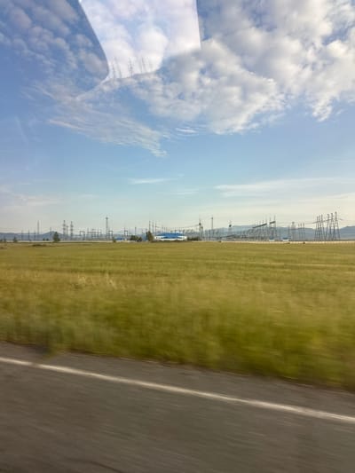 The image displays a scene captured from a moving vehicle, looking towards a vast, grassy field. A portion of the road is visible on the right side of the frame, with a distinct white line marking its edge. Beyond the field, a power substation with numerous tall metal towers and electrical wires stretches across the landscape. In the distance, mountains can be seen, partially obscured by the power infrastructure. The sky is a clear blue with scattered white clouds, suggesting fair weather. The lighting indicates it is daytime. The blurriness of the image suggests the photo was taken while in motion, likely from a car or train. There are no people or animals visible in the frame. The overall impression is of an open, rural or semi-industrial landscape under a bright sky. There is no visible text in the image.