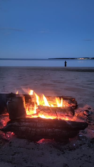 An evening scene on a sandy beach shows a campfire with burning logs in the foreground. A metal pot is placed on the left side of the fire. The calm sea stretches out towards the horizon, reflecting the dusky blue sky. A lone figure stands silhouetted on the beach in the distance. In the background, along the far shore, lights from a settlement are visible. The location is identified as Zaostrov'e, Russia.