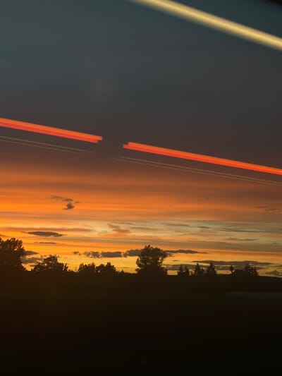 A sunset is captured over a silhouetted landscape of trees and fields. The sky is a vibrant gradient of orange, yellow, and dark blue, with streaks of light that appear to be from vehicle headlights or contrails. The clouds are scattered throughout the colorful sky, some appearing dark and others illuminated by the setting sun. The lower portion of the image is dominated by a dark silhouette of trees and vegetation, suggesting a rural or suburban area. The scene is likely captured from a moving vehicle due to the light streaks. The image does not contain any identifiable landmarks or features that would place it specifically in Baiersdorf, Germany.