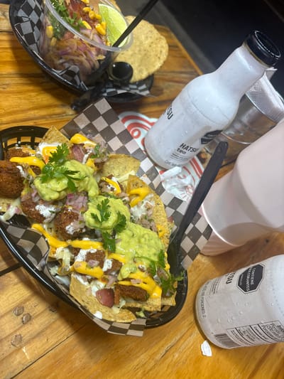 The image depicts a table setting with food and beverages. On the wooden surface, there is a black basket filled with nachos topped with guacamole, chopped onions, what appears to be fried protein pieces, and a yellow sauce, possibly cheese. Next to it, there is a smaller basket with what seems to be a salad or a corn salsa. Two bottles labeled "HATSU tea" are also visible, indicating a potential location in a region where this brand is distributed. The bottles are covered in condensation, suggesting they are cold. Based on the ingredients and presentation, the scene likely represents a dining experience at a restaurant or food establishment. As the prompt specifies that this picture was taken in Envigado, Colombia, it is likely that the photograph depicts food and drinks offered at a local establishment there.