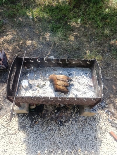 The image shows a portable metal barbecue grill filled with ash and a piece of wood, situated on a gravel surface. The grill is elevated by two concrete blocks. A green bottle with a label partially visible that says "BABA" is placed underneath the grill. A white crumpled paper and cigarette butts are also visible in the ash. The surrounding environment consists of dry grass and dirt, with shadows from trees indicating an outdoor setting. The scene appears to be in a natural or semi-natural area, likely a park or backyard, located in Dumeni, Moldova. There are no people visible in the image.
