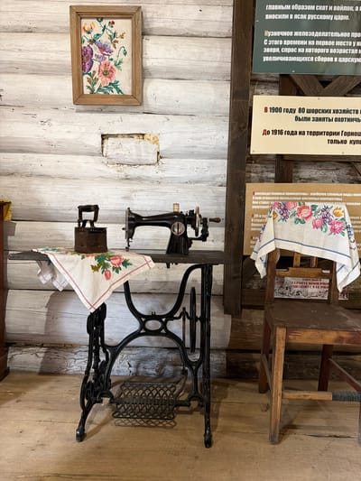 The image depicts an indoor scene, possibly a museum exhibit, set against a backdrop of a log wall. In the foreground, a vintage black sewing machine with an ornate cast-iron base and pedal mechanism stands on a dark wooden surface. To the left of the sewing machine, an antique cast-iron flatiron rests on a white embroidered cloth featuring floral motifs of pink, purple, and green flowers. Above the sewing machine, a framed embroidered artwork of similar floral patterns hangs on the wall. To the right, a wooden chair is positioned with another embroidered cloth draped over its back, also displaying a floral design. Several informational signs with text in Russian are mounted on the log wall behind the exhibit. The floor is made of light-colored wooden planks. The overall impression is one of a historical display showcasing domestic life and crafts. The location is identified as Ust'-Kabyrza, Russia, suggesting this scene is part of a local museum or historical site within that city. There are no people present in the image.