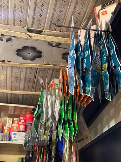This media file shows a close-up, horizontal shot of various small, packaged items hanging vertically. The items appear to be electronic accessories, such as charging cables or earbuds, with colorful packaging in shades of blue, green, and orange. Some of the packaging has visible text, including what appears to be brand names like "JBL" and "BOSS". The items are arranged in multiple rows, creating a dense display.
In the background, the scene includes patterned tiled walls or surfaces with a diamond and swirl design. To the right, there are shelves filled with what seem to be boxes of light bulbs, some red and white, with visible text in English and possibly other languages. A black electronic device, possibly a speaker, is also visible to the far right. The lighting suggests an indoor environment, possibly a shop or market stall. The overall impression is a retail setting stocked with electronic goods and lighting products. The image appears to be taken in Barakin, Nigeria.
