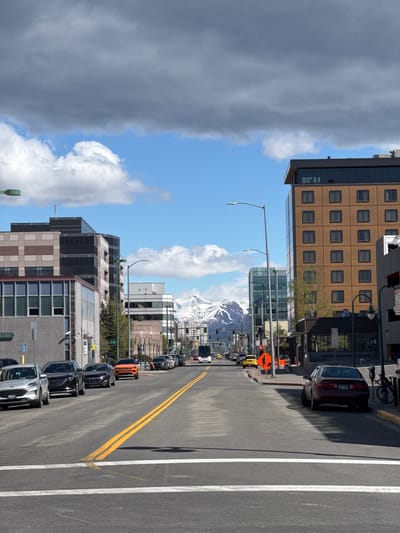 The image captures a street view in Anchorage, Alaska, United States. The scene is taken from the side of the road, showing cars parked along the curb. An orange car and several other vehicles are visible, parked in a line. The road features double yellow lines marking the center and a white line along the edge. The background includes buildings of varying heights and architectural styles. A mountain range, partially obscured by clouds, can be seen in the distance under a clear, blue sky. The sky also has some gray clouds. Overall, the image presents an urban setting with a mountainous backdrop.