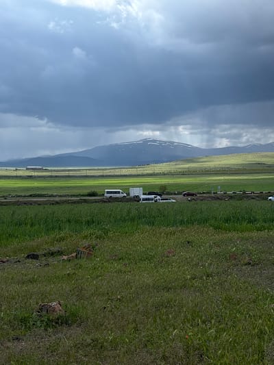 The image depicts a landscape scene in Tlik, Armenia. A mountain with snow patches is visible against a cloudy sky. In the foreground, there is a road with several parked white vans. Adjacent to the road are green fields. The overall impression is a rural or semi-rural environment.
