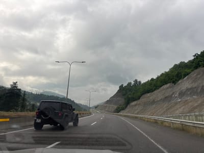 This media file depicts a view from inside a vehicle traveling on a highway in Tsakva, Georgia. The dominant subject is a black Jeep Wrangler in the foreground, seen from its rear quarter panel. The vehicle has a spare tire mounted on the back and a license plate that reads "WO - 888-OW". The highway has multiple lanes and is wet, suggesting recent rain. The scene is set in a mountainous, green landscape under a cloudy, overcast sky. On the right side of the highway, a steep, terraced slope is visible, reinforced with concrete and covered with greenery and trees. On the left side, there is a highway barrier and more trees leading up to rolling hills. Several streetlights are visible along the road. The overall mood is somber, likely due to the weather and the grey sky. The time of day appears to be daytime, given the visibility, but the heavy cloud cover obscures any direct sunlight.