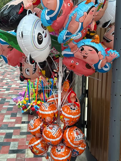 An outdoor street stall displays a variety of colorful balloons and small toys. In the foreground, a cluster of orange balloons with white stripes and images of cartoon characters are attached to plastic sticks. Some of these balloons have text in what appears to be English, including "Anjanli bol" and "TOY SURPRISE." Behind these, a large white balloon shaped like a panda's face is visible, along with other character-shaped balloons in shades of blue, pink, and black. A rack of colorful plastic toys, including what look like mini bicycles and characters with wheels, is situated behind the balloons. The ground is paved with interlocking rectangular tiles in shades of red and grey. The background includes a wooden door or wall on the right. The scene appears to be a commercial setting, possibly a market or a street vendor's stall, in the city of Manas, Kyrgyzstan. There are no people clearly visible in the image.