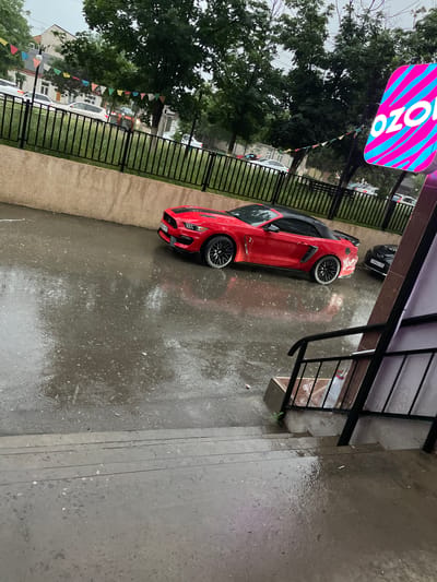 A red convertible Mustang is parked on a wet, paved surface, possibly a street or parking lot, in Buynaksk, Russia. The car is facing towards the right side of the frame. It is a cloudy day, with rain having recently fallen, evidenced by the wetness and reflections on the ground. In the background, there are trees, a black metal fence, and buildings. A brightly colored sign with the word "OZON" in large white letters, set against a backdrop of pink and blue diagonal stripes, is visible on the right side of the image. There are also several other cars parked in the background. A black sedan is parked behind the red Mustang. In the foreground, a concrete staircase leads down to the paved area.