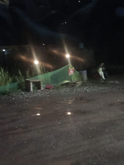 The image depicts an outdoor scene at night in Kathwaharn, India. A green tent is erected, and a person is partially visible sitting near it. There is a large cylindrical object, possibly a barrel, next to the tent. In the foreground, the ground appears to be wet and reflective, suggesting recent rain or puddles. The background consists of dark, out-of-focus structures and vegetation. Several lights are visible, some appearing as bright orbs and others as streaks from being out of focus. There are also some small, reflective objects scattered on the ground.