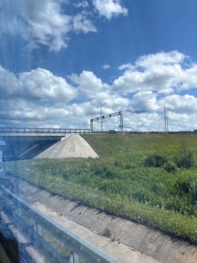 The image depicts a view from a moving vehicle on a sunny day, likely on a highway in the vicinity of Myasnoi Bor, Russia. The foreground shows a concrete drainage ditch bordered by a metal guardrail, with some light smoke or haze obscuring the view to the left. Beyond this, a grassy embankment leads up to a highway overpass. The overpass is characterized by a concrete structure with a sloped, paved approach and a metal railing. Overhead, a gantry with traffic signs and streetlights spans the highway. The sky is a vibrant blue, dotted with fluffy white cumulus clouds. The environment is predominantly green, with lush grass and scattered wildflowers on the embankment. The overall scene suggests travel through a semi-rural or highway landscape under a clear, partly cloudy sky.