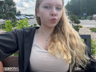 A young woman with long blonde hair and fair skin is taking a selfie outdoors. She is wearing a beige top and a black jacket. Her lips are pursed, and she has a necklace with a small pendant. Behind her, a parking lot is visible with several cars, including a white Volvo. In the background, there are trees and green trash bins. The setting appears to be a public area, possibly a rest stop or a parking lot near a natural area, in or near Камянюцкі сельскі Савет, Belarus. The weather seems to be overcast with some clouds.