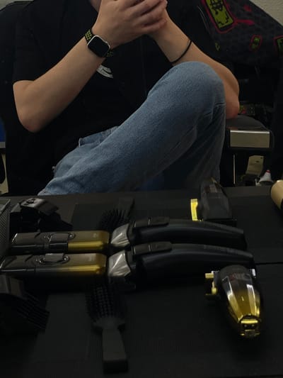 The image shows a close-up of a person's lower legs and arms, with a set of hair clippers and brushes in the background. The person is wearing blue denim jeans and a black shirt, with a smartwatch on their left wrist. The hair clippers are black and gold, and the brushes are black. The scene appears to be indoors, possibly in a salon or a home grooming area. The lighting is dim, suggesting it might be evening or the room is not well-lit. The person's posture suggests they are sitting or resting. There is no visible text in the image.