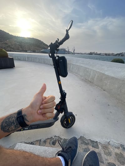 The image shows a person's arm and hand giving a thumbs-up gesture, positioned next to an electric scooter. The scooter is black with yellow accents on the wheel. A black bag is attached to the handlebars, and a mirror is visible on the left side of the handlebars. The person is wearing a black watch on their wrist and has tattoos on their forearm.
The scene appears to be outdoors, with a concrete walkway or promenade. In the background, there is a body of water, likely the sea, and a cityscape with buildings and cranes along the coast. Rocky hills or mountains are visible in the far distance, with the sun shining brightly. The sky is bright and somewhat cloudy, suggesting it is daytime, likely late afternoon or early evening given the low angle of the sun. The location is identified as Cartagena, Spain. The overall mood seems positive and relaxed, implied by the thumbs-up gesture and the scenic background. There is no visible text in the image.