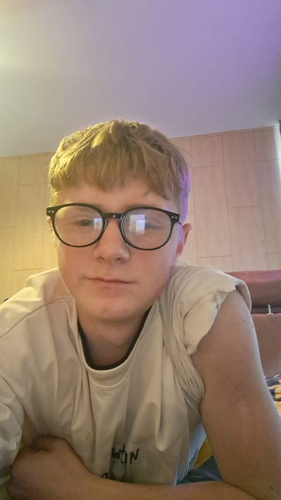 The image shows a close-up, rotated portrait of a young person wearing glasses and a light-colored t-shirt. The person has fair skin, short blonde hair, and is looking towards the camera. The glasses have thick black frames and round lenses. A portion of the person's arm and shoulder is visible on the left side of the image. The background appears to be a tiled wall and a wall illuminated with purple light, possibly from an LED strip. The lighting is warm and suggests an indoor setting. The overall mood is casual and relaxed. There is visible text on the t-shirt, though it is partially obscured and difficult to read clearly, appearing to be the letters "NOW". The image appears to have been taken indoors.