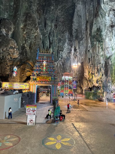 This image captures a vibrant Hindu temple complex nestled inside a vast, natural cave. The cave walls, dominating the left side and top of the frame, are rugged and textured, displaying various shades of grey, brown, and hints of green from mineral deposits. Artificial lighting illuminates sections of the cavern, highlighting its grand scale and intricate geological formations.

The focal point is a colorful, multi-tiered temple structure, characteristic of South Indian (Gopuram) architecture, situated towards the center of the cave. It is richly adorned with numerous intricate sculptures and brightly painted details. Surrounding the main structure are various smaller shrines and decorated alcoves. A large yellow banner on the right side of the temple clearly identifies it as "ஸ்ரீ வேலாயுதர் கோவில் (மூலஸ்தானம்) SRI VELAYUTHAR TEMPLE (MAIN TEMPLE)". An illuminated trident-like symbol is also visible on scaffolding near the temple.

The ground area, occupying the right and foreground, is a paved surface decorated with large yellow and red floral patterns. Several people are present, either walking or standing, observing the temple or moving within the cavern. One person in a light vest stands near a prominent white sign. The sign provides guidelines for visitors, stating "YOU ARE ENTERING," "NO SHOES," and indicating a dress code, no pets, and no food/drinks. It also contains text in Malay: "ANDA MEMASUKI KE KAWASA PUKAIPIADKAIR," likely a warning against smoking.

The scene depicts a sacred site where devotees and visitors can explore the temple within the awe-inspiring natural setting of the cave. The artificial lighting creates a dramatic ambiance, suggesting a perpetually lit interior regardless of the time of day outside. The presence of multi-language signage and the temple's style align with locations like the Batu Caves in Malaysia.