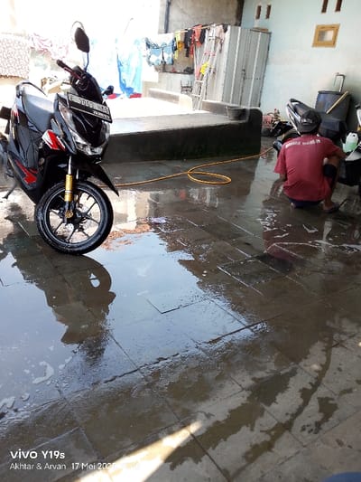 The image shows a wet patio in Gegesik, Indonesia, at the coordinates -6.533 latitude and 108.432 longitude. There is a black motorcycle parked on the left side of the frame. A person is sitting on the floor to the right of the motorcycle, cleaning the motorcycle. The floor is wet with soapy water and there is a yellow hose nearby.  The surrounding environment is a patio with a wall and a metal gate on the left side and a building with a window on the right side. The patio appears to be constructed of stone pavers.
