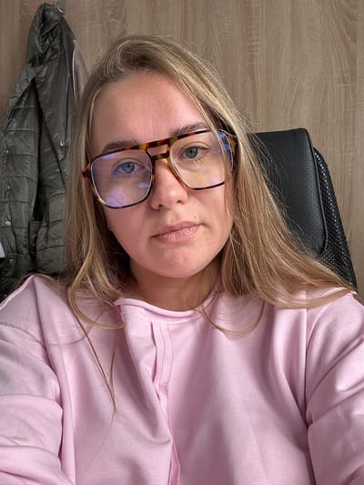 A person with light skin, blonde hair, and blue eyes is visible in a selfie-style photograph. The person is wearing eyeglasses with a tortoiseshell pattern frame and a light pink hooded sweatshirt. They are reclining against a black, perforated material that appears to be the headrest of a chair. A folded gray jacket with a quilted pattern is beneath the person's head. The background consists of a wooden-patterned surface, which may be a table or floor. No indication shows Ural'skoe, Russia.
