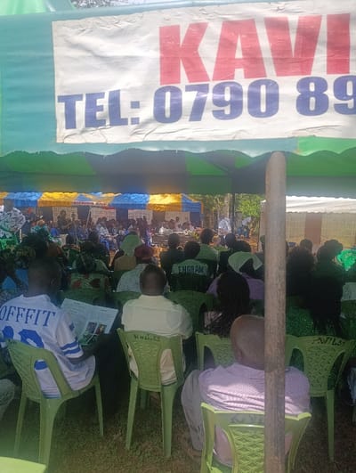 The image depicts an outdoor gathering under multiple large tents in Muthetheni ward, Kenya. In the immediate foreground, a white banner with red text "KAVI" and blue text "TEL: 0790 89" is stretched across the underside of a green and blue striped tent. Below this banner, numerous individuals are seated on green plastic chairs, facing away from the camera towards the front of the gathering.
Essential details about people and subjects:
* In the lower-left foreground, a male subject wears a white jersey with blue numbering "93" and the word "OFFFIT" across the shoulders. He is holding and looking at an open booklet or magazine.
* Next to him, another male subject wears a light-colored long-sleeved shirt.
* Further back, a subject wears a green top with "ENGRAM" written in white on the back.
* To the right, partially obscured by a wooden pole, a male subject is visible from the back, wearing a light purple checkered shirt.
* The seated individuals are predominantly adults, dressed in various casual attire. Many are facing forward, suggesting an audience for a speaker or presentation.
Essential details about objects:
* The primary objects are numerous light green plastic chairs, some bearing markings such as "KVA.33" on their backs.
* Multiple tents are visible: a prominent green and blue/yellow striped tent in the foreground, and other blue and yellow striped tents further back.
* The "KAVI" banner, partially obscuring a phone number, is a key object under the foreground tent.
* A thick, unpainted wooden pole stands vertically in the right-center foreground, slightly obstructing the view of the attendees.
* The subject on the far left holds a small, open publication.
Essential details about actions and interactions:
* The main action is a large group of people seated and observing an unseen focal point.
* The individual on the left is engaged in reading.
* The overall arrangement suggests an organized public or community event.
Essential details about the environment:
* The ground is an unpaved surface, likely dirt or sparse grass, visible beneath the chairs.
* The scene is outdoors, under natural daylight conditions, with shadows indicating bright sun.
* The background shows more tents and possibly some distant foliage, indicating a semi-rural or open-area setting within the Muthetheni ward.