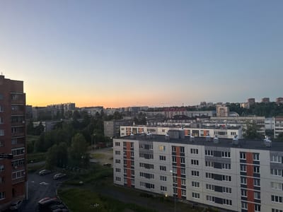 The image captures a cityscape of Kondopoga, Russia, during dusk. In the foreground, a tall, brick apartment building is visible on the left, with multiple windows and balconies. To the right, a modern apartment building with a white, grey, and orange facade stands prominently. Several cars are parked in the lot between the buildings, and streetlights illuminate the scene. Beyond these structures, a panorama of residential buildings, ranging in height and architectural style, extends into the distance. Lush green trees are interspersed throughout the urban landscape, and the sky transitions from a pale orange near the horizon to a soft blue overhead, indicating the twilight hour. The overall impression is of a typical residential district in a Russian city as the day concludes.