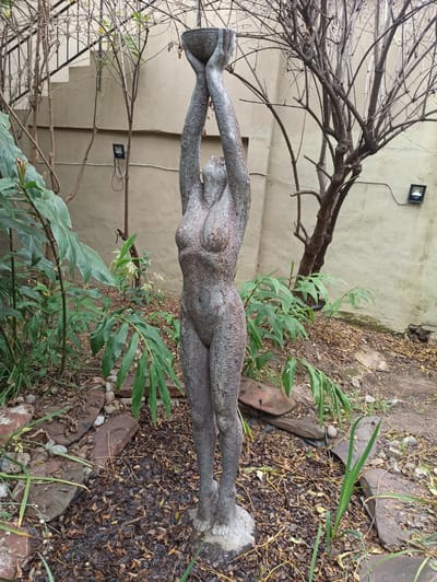 This is a full-shot, eye-level, outdoor photograph of a weathered, stone statue of a female figure with arms raised, holding a bowl. The statue is centrally positioned in a garden. The figure is anatomically detailed, with a rough, textured surface. The background consists of a light-colored, stucco wall with two black outdoor light fixtures mounted on it. To the left of the statue, there is a metal staircase with black railings, partially obscured by lush green foliage. Bare tree branches with no leaves are visible behind the statue and to the right. The ground is covered with dry leaves, small rocks, and some larger flat stones that appear to form a rudimentary pathway. There are also some green plants and saplings growing in the foreground and around the base of the statue. The lighting suggests an overcast day. The location is the garden of a residential property in Randburg, South Africa.