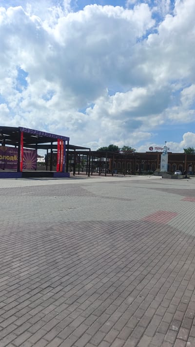 The image depicts an outdoor plaza with a paved walkway in the foreground. In the middle ground, there is a wooden stage structure with a canopy, decorated with banners. One banner features text in Cyrillic that appears to say "Молодёжь" (Youth), followed by "Мы - энергия года!" (We are the energy of the year!). Other banners are red and blue with Cyrillic text that appears to read "ЕДИНСТВО" (Unity), "ГОРДОСТЬ" (Pride), and "МЕЧТА" (Dream). Beyond the stage, there is a long, brick building with arched windows and a covered walkway in front of it. A sign with a red "5" and Cyrillic text reading "Пятерка" (Five) is visible on the building, possibly indicating a store or business. To the right of the building, a sculpture or monument stands in front of it, featuring a star on top. Several cars are parked near the building. The sky is filled with large, white cumulus clouds against a blue background, suggesting it is daytime with fair weather. A few pigeons are scattered across the plaza. The overall scene suggests a public gathering space, possibly a town square or park, set up for an event.