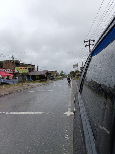 A street view from a car window shows a wet road with white lane markings. Buildings line the street, including one with a "Bakso MX" sign. Power lines stretch across the sky. Two motorcyclists are further down the road. The day is overcast and grey.
