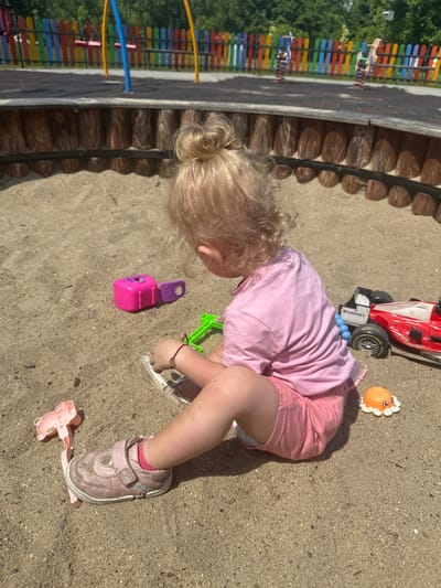 A young child with blonde hair pulled into a bun is sitting in a sandbox, facing away from the camera. The child is wearing a pink t-shirt and pink shorts, and pink shoes. The child's left hand is in the sand, and they appear to be playing with sand. Several toys are scattered in the sandbox around the child, including a pink toy, a green shovel, a red and black toy car, and a small orange octopus toy. In the background, a wooden border surrounds the sandbox, and beyond that, a colorful fence and playground equipment are visible, suggesting the location is a public playground. The overall setting is outdoors during the daytime, with bright sunlight casting shadows. The scene is consistent with a playground in Donchovci, Bulgaria, given the typical features of such facilities.