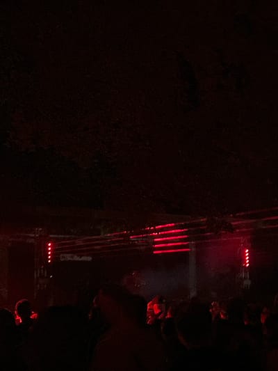 A low-angle, dimly lit photograph captures a crowd of people gathered in what appears to be a concert or performance venue in Viana do Castelo, Portugal. Red lights illuminate the scene, casting a crimson glow on the dark environment and the silhouettes of individuals. In the background, horizontal structures with multiple red lights are visible, suggesting stage lighting or a performance setup. The crowd is densely packed, with many figures appearing as dark silhouettes against the red illumination. The overall atmosphere is dark and energetic, characteristic of a live event.