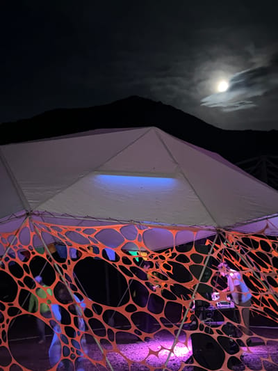 This is a night photograph taken in Kashka Suu, Kyrgyzstan, featuring a temporary outdoor structure resembling a large tent or canopy. The structure is illuminated with vibrant neon lights, casting a purple and blue glow. An orange, web-like decorative netting hangs from the tent's frame, creating a layered effect and partially obscuring the view of people inside. Within the tent, at least two individuals are visible, one appearing to be operating DJ equipment. The background is dominated by the dark silhouette of mountains under a moonlit sky, with the moon partially obscured by clouds. The ground appears to be covered in gravel or small stones, also illuminated by the colored lights.